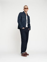 Veste en denim foncé avec fermeture à boutons, assortie à un t-shirt blanc et à un jean foncé. Chaussures brunes avec lacets et semelles en caoutchouc. Lunettes de soleil teintées orange.
