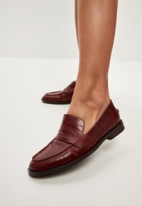 Burgunderfarbene Lederloafers mit glatter Oberfläche, ausgestattet mit einem strukturierten Band an der Vorderseite und einer niedrigen schwarzen Sohle.