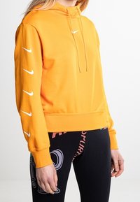 Oranžová mikina Nike s kapucí, přední kapsou, stahovací šňůrou a bílým logem; po rukávech jsou bílé akcenty ve tvaru swoosh. Kombinována s černými legínami.