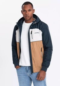 Herren Farbblockjacke mit navy, beige und weißen Paneelen. Wasserabweisendes Material, mit einer Reißverschlusstasche auf der Brust und verstellbarem Kapuzen. 