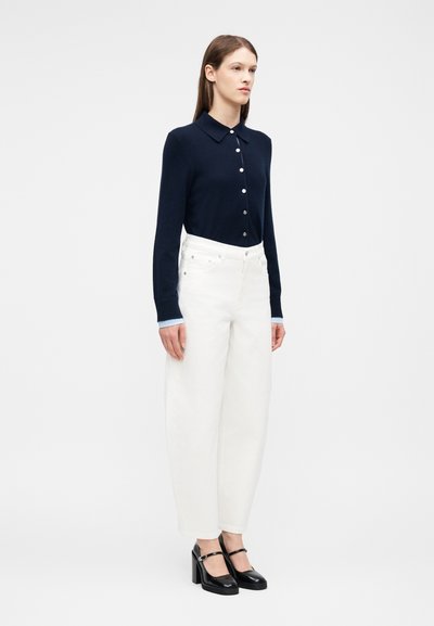 Γυναίκα που στέκεται φορώντας μπλε navy πουκάμισο με μακριά μανίκια και κουμπιά, λευκό παντελόνι με ψηλή μέση και φαρδύ πόδι, και μαύρες γόβες Mary Jane.