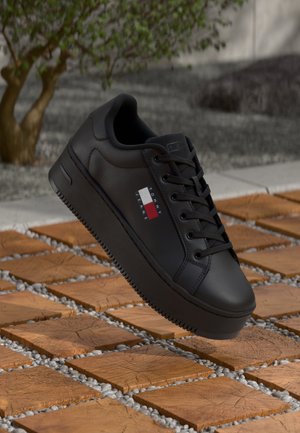 Sort Tommy Jeans sneaker med tyk sål og snørebånd, ophængt over træterninger og små grå sten udendørs.