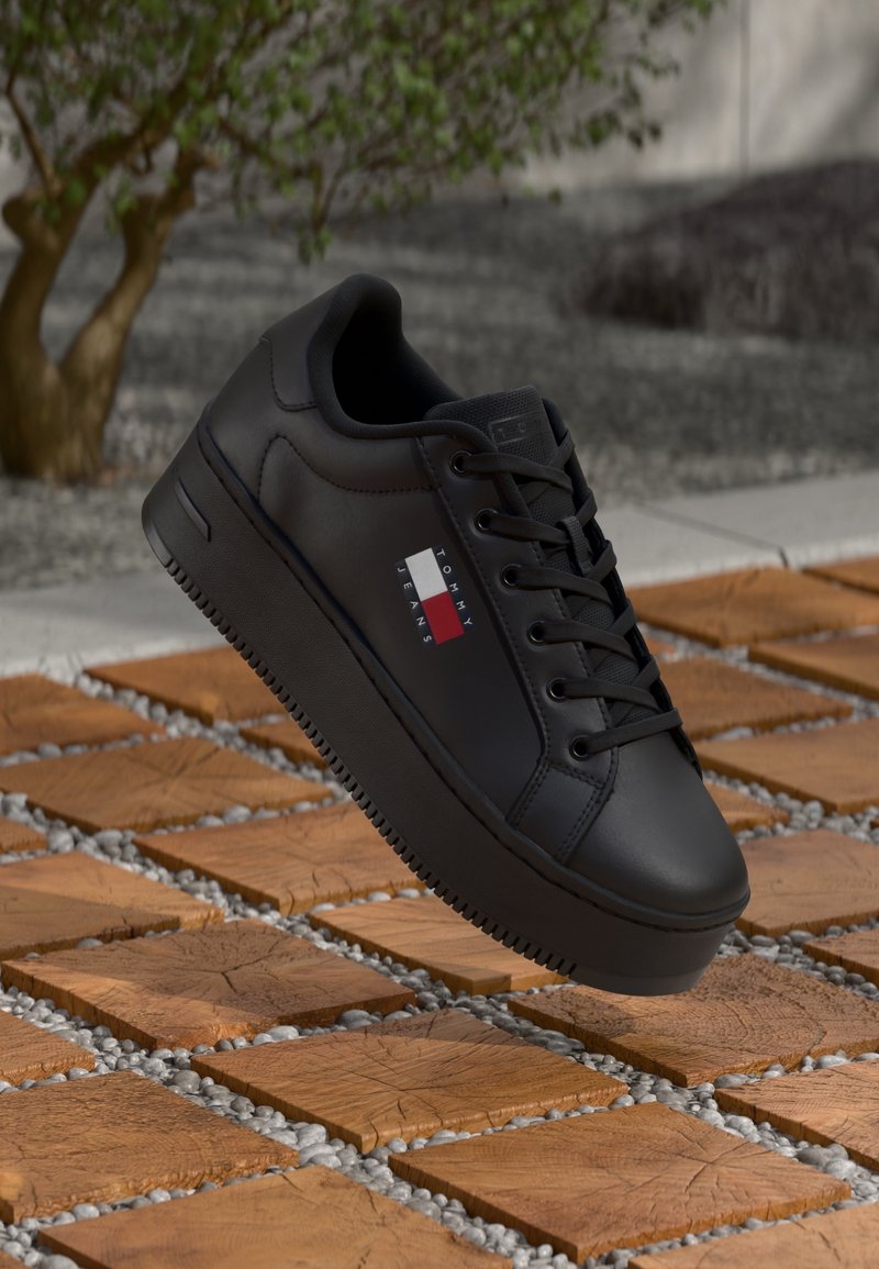 Baskets noires Tommy Jeans avec semelle épaisse et lacets, suspendues au-dessus de carreaux en bois en forme de cube et de petits cailloux gris en extérieur.