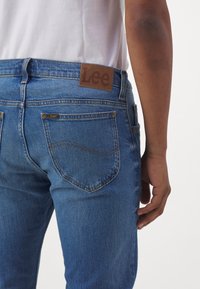 Blaue Denim-Jeans mit einem braunen Lederetikett am Bund, fünf Taschen und gesteppten Akzenten. Rückansicht, Modell trägt ein weißes Hemd.