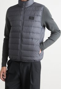 Szara pikowana kamizelka z wysokim kołnierzem, zapięciem na zamek i naszywką z logo CK. Noszona na ciemnej bluzce z długim rękawem, o pikowanej teksturze.