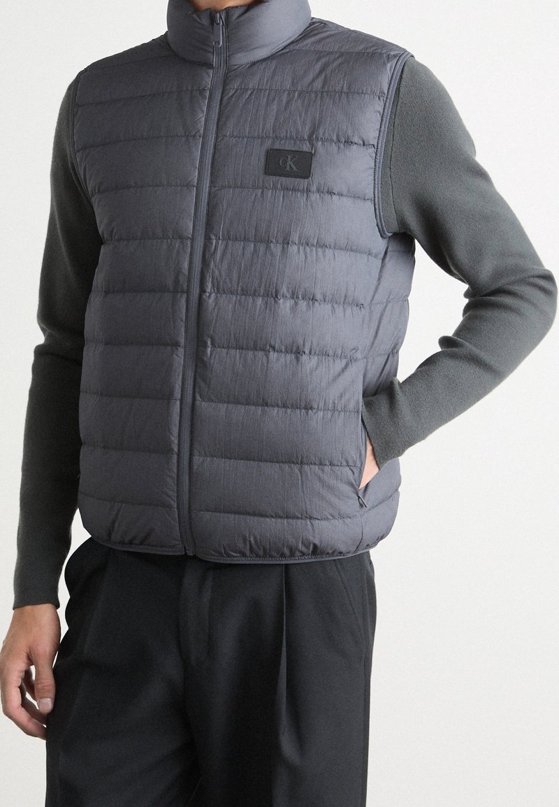 Szara pikowana kamizelka z wysokim kołnierzem, zapięciem na zamek i naszywką z logo CK. Noszona na ciemnej bluzce z długim rękawem, o pikowanej teksturze.