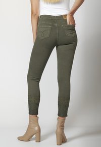 Mujer vestida con jeans ajustados de color verde oliva con bordados decorativos en los tobillos y botas de tacón beige hasta el tobillo, de pie contra un fondo liso.