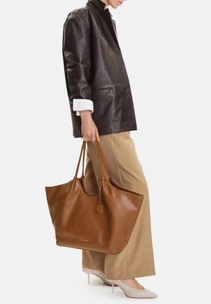 Persona che indossa una giacca di pelle scura e pantaloni beige, tiene una grande borsa a tracolla in pelle marrone, indossando tacchi bianchi a punta.
