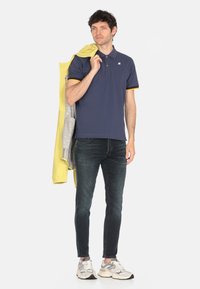 Polo navy con dettagli gialli, abbinato a jeans scuri. Il modello tiene una giacca gialla sulle spalle, indossando sneakers grigie e bianche.
