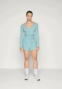 Kvinde står imod hvid baggrund iført en lys blå langærmet omslagstop og matchende shorts med hvide sneakers og sokker.