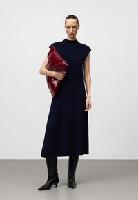 Vestito senza maniche blu navy con scollo alto, abbinato a una borsa a tracolla rossa lucida. La modella indossa stivaletti neri a punta.