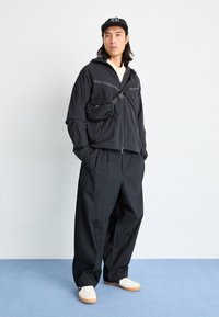 Μαύρο μπουφάν με φερμουάρ και ανακλαστικές ρίγες, συνδυασμένο με χαλαρές μαύρες παντελόνες και λευκά αθλητικά παπούτσια. Η μαύρη τσάντα χιαστί προσθέτει λεπτομέρεια.