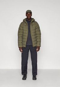 Νεαρός άντρας φορώντας μια ελαιόχρωμη μπουφάν, μαύρη ζακέτα με φερμουάρ, μαύρο παντελόνι cargo, μαύρα παπούτσια και ελαιόχρωμο καπέλο, που στέκεται μπροστά σε λευκό φόντο.