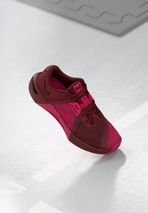 Rode en donkerbruine sportschoen met zwart logo, mesh textuur en veters, zwevend boven een grijze reflecterende vloer met witte lijnen.