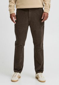 Pantaloni di velluto a coste marroni con tasche laterali, vestibilità affusolata, texture a righe e cuciture rinforzate. Indossati con sneakers bianche con strisce marroni.