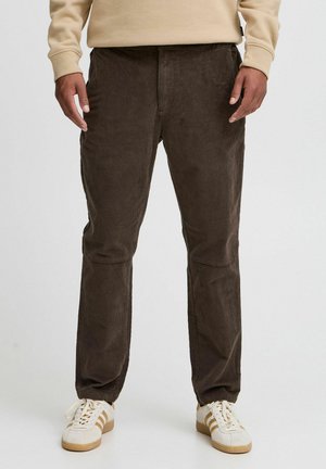 Pantaloni di velluto a coste marroni con tasche laterali, vestibilità affusolata, texture a righe e cuciture rinforzate. Indossati con sneakers bianche con strisce marroni.