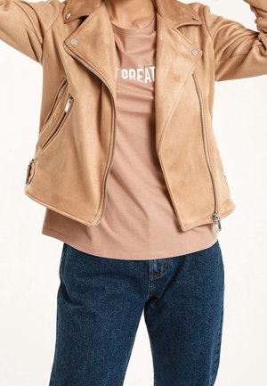 Henkilö pukeutuneena beigeen mokkatakkiin, jonka alla vaaleanruskea paita, jossa osittainen valkoinen teksti, siniset farkut ja kädet kohotettuina poseeraamassa.