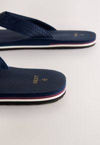 Marineblaue Flip-Flops mit strukturierten Fußbetten, flachen Riemen und einer mehrfarbig gestreiften Sohle. Mit in Gold geprägtem Branding auf dem Fußbett.