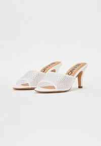 Zapatillas de tacón abiertas en la parte delantera, de color blanco, con una parte superior de malla que presenta patrones de diamante, plantilla marrón y un delgado tacón de aguja. Nombre de la marca "BIANCO" visible.
