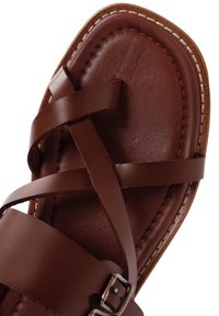 Sandale en cuir marron avec des brides croisées, semelle plate et détails de couture légers. Dispose d'une boucle en argent pour un maintien sécurisé.