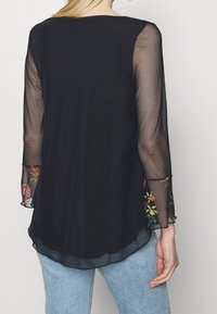 Kvinde iført sort gennemsigtig langærmet bluse med blomsterbroderede manchetter, parret med lyseblå jeans, vist fra ryggen.
