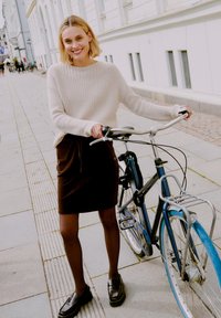 Pull en laine crème, jupe en velours côtelé marron, collants noirs et mocassins, se tenant à côté d'un vélo bleu sur une rue piétonne.