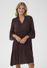 Abito marrone scuro, con lunghezza al ginocchio, scollatura a V, maniche a 3/4 svasate e design a strati. Realizzato in un tessuto leggero e testurizzato.