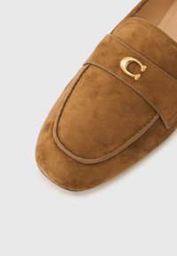 Καφέ μοκασίνια σουέντ με στρογγυλεμένη μύτη, με διακοσμητικό χρυσό γράμμα "C" και ραμμένη λεπτομέρεια στο επάνω μέρος. Ομαλή υφή.