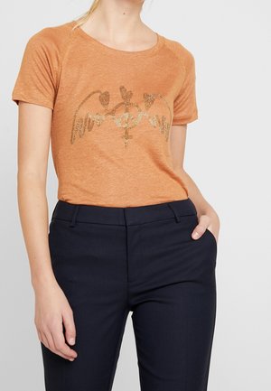 Kobieta ubrana w pomarańską koszulkę z krótkim rękawem z złotymi wzorami pokoju i serca, w połączeniu z granatowymi spodniami z wysokim stanem.