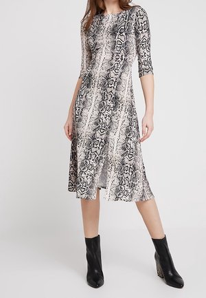 Femme portant une robe mi-longue à imprimé serpent avec des manches trois-quarts et des bottines noires, debout devant un fond uni.