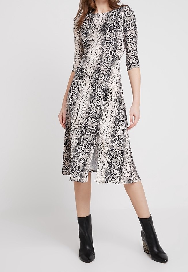 Femme portant une robe mi-longue à imprimé serpent avec des manches trois-quarts et des bottines noires, debout devant un fond uni.