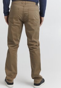 Braune Baumwollhosen mit lässiger Passform, ausgestattet mit zwei Gesäßtaschen und einem Etikett, kombiniert mit schwarzen Sneakers. Sichtbares Nähdetail.