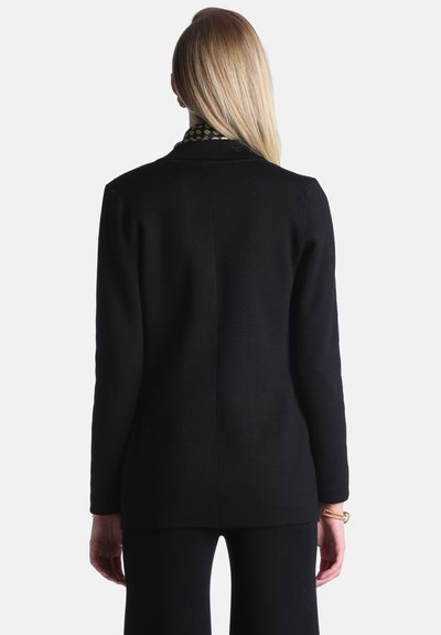 Femme blonde vue de dos portant un blazer noir et un pantalon noir assorti avec des bracelets dorés à son poignet droit.