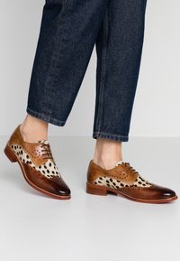 Chaussures en cuir marron avec des accents en cuir de vache tacheté, un design brogue et un bout arrondi. Portées avec un jean foncé, mettant en avant les lacets et les perforations.