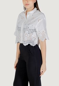 Camicia bianca a maniche corte con motivi floreali intagliati e orlo a ondine. Presenta una chiusura con bottoni sul davanti e colletto, abbinata a pantaloni neri.