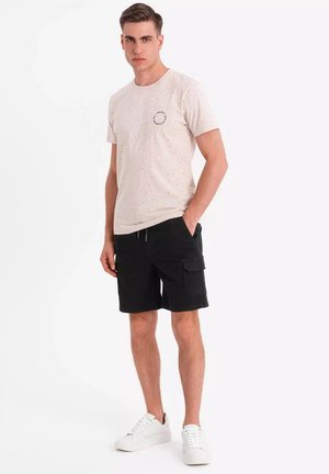 Camiseta de manga corta en color beige claro con diseño de manchas y un logo en el pecho, combinada con pantalones cortos cargo negros y zapatillas blancas.