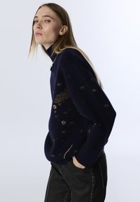 Navy knitted sweater with a high collar, featuring gold star patterns and a loose fit. Set against a light background, paired with black pants.