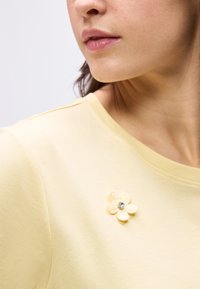 Prizor od blizu osebe, ki nosi svetlo rumeno srajco z majhnim tkaninskim cvetom, okrašenim s prozornim kamnom na izrezu.