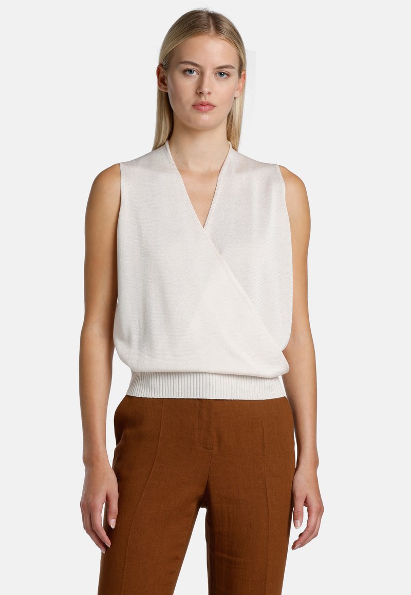 Top sin mangas en crema con un escote en V profundo, diseño envolvente y una cinturilla fruncida. Textura suave con un ligero brillo, combinado con pantalones marrones.