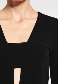 Czarna bluzka z długim rękawem, głębokim dekoltem w kształcie litery V i wycięciem na klatce piersiowej, wykonana z gładkiego materiału i o dopasowanym kroju.