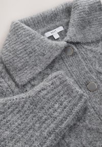 Maglione grigio a maglia lavorata con collo a camicia e due bottoni neri. Il tessuto sembra morbido, con un motivo a coste e una lieve peluria.
