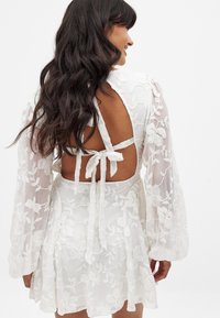 Vestido blanco con bordados florales, mangas largas transparentes, espalda descubierta y cierre con lazo, que presenta una falda acampanada y detalles intrincados.