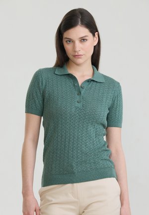 Frau trägt ein grünes, kurzärmeliges Strick-Poloshirt mit Knopfleiste und beigen Hosen, steht vor einem schlichten hellen Hintergrund.