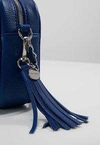 Borsa in pelle blu con superficie testurizzata, zip e accessori in argento, presenta un accento in pelle con nappina. Etichetta circolare del marchio attaccata.