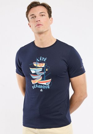 Homme portant un t-shirt bleu marine avec un graphisme de petits bateaux, des bouées de sauvetage et le texte français "L'été Débarque" sur le devant.