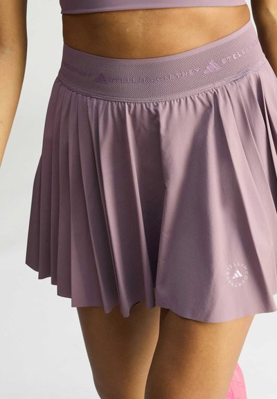 Woman wearing a lavender pleated athletic skirt with perforated waistband and logo near hem, standing against a plain background.