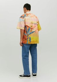 Camisetta a maniche corte multicolore con stampa tropicale su tessuto leggero, dal taglio rilassato, abbinata a jeans denim blu e sneakers nere.