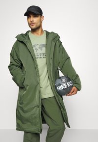 Grüner gepolsterter langer Mantel, passendes grünes T-Shirt mit Grafik, grüne Hose, schwarze Mütze, hält einen schwarzen Nike F.C. Fußball, glatte Textur.