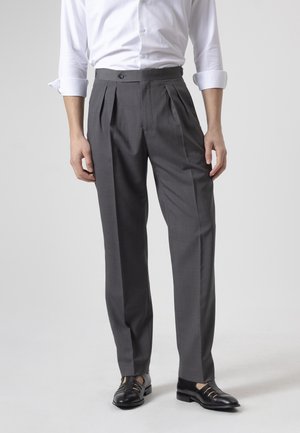 Hombre con pantalones de vestir grises de pinzas, camisa blanca de manga larga con puños remangados y sandalias de cuero negras sobre un fondo liso.