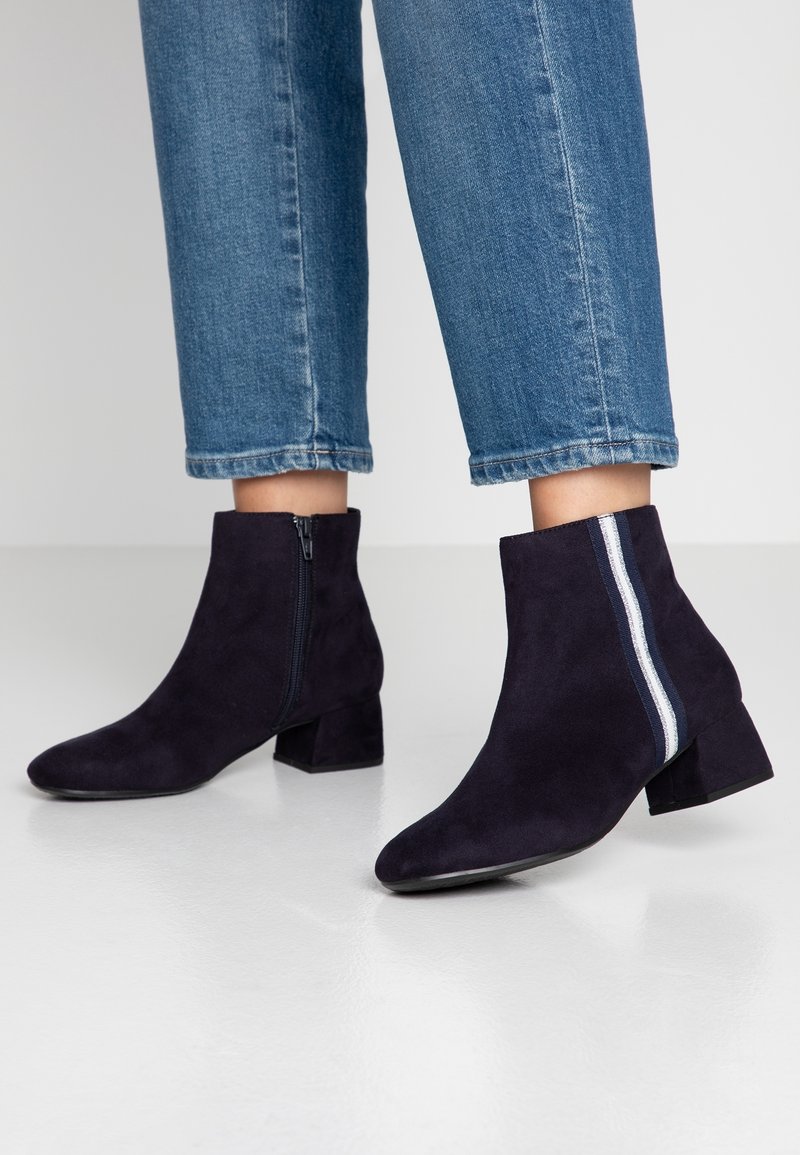 Bottes en daim foncé longueur cheville avec talons carrés, fermeture éclair latérale et rayures blanches et bleues, portées avec un jean court bleu.